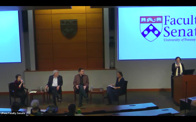 Three faculty members sit at the front of an auditorium having a discussion. A fourth stands at an adjacent lectern.